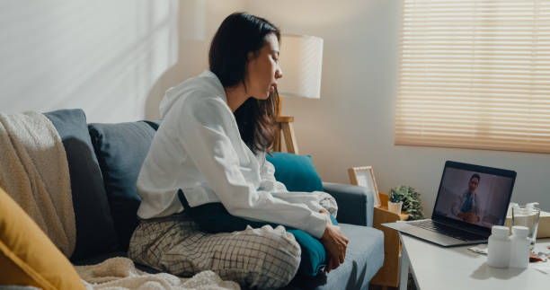 노트북을 사용하는 아픈 젊은 아시아 여성은 집에있는 거실의 소파에 앉아있는 의사와 이야기합니다. - pill men home interior one person 뉴스 사진 이미지
