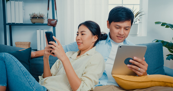 Young Asian married couple with casual sitting on couch use smartphone and tablet shopping online and watch online movie in living room at home.