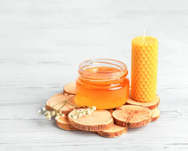 Glass jar with honey, beeswax candle, white wildflower on a wooden stand on a white wooden background, front view, close-up, copy space. Alternative medicine, healthy eating, vegetarianism.