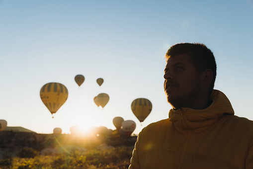 One male explorer in warm jacket walking on the hill during scenic pink sunrise with hundreds of air balloons on the sky in Turkey