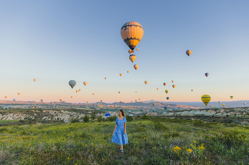 Young female with long hair in a blue dress contemplating the beautiful dawn above the picturesque valley with hundreds of hot air balloons in Turkey