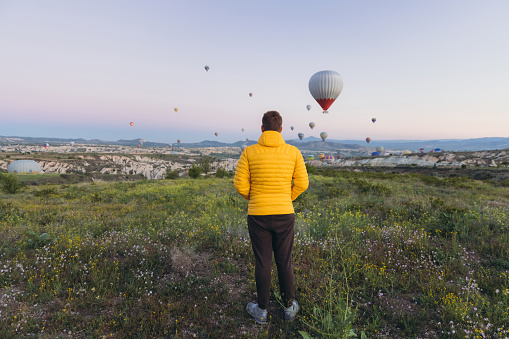 One male explorer in warm jacket walking on the hill during scenic pink sunrise with hundreds of air balloons on the sky in Turkey