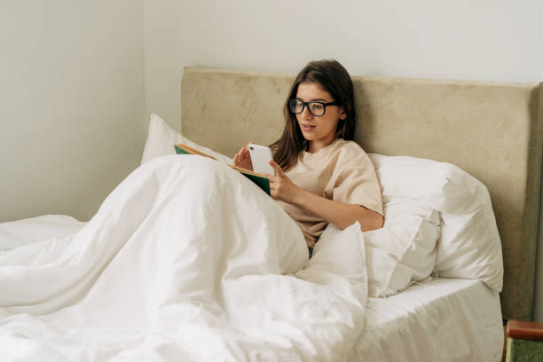 istock Female pretty student reading a book and taking notes on mobile phone while lying in bed 1404231159