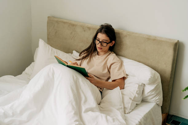 istock Young brunette female student in eyeglasses  reading a book and taking notes while lying in her bed 1404231160