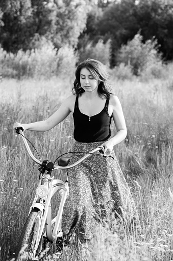 sad brunette girl cyclist walking with bike at sunny meadow, monochrome