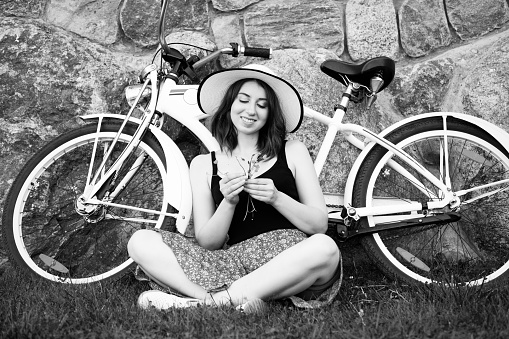 pretty brunette girl with bike near wall resting, sitting in grass with flowers, monochrome