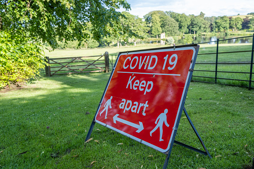 A red sign warning about the Coronavirus pandemic and social distancing in a field at an outdoor event