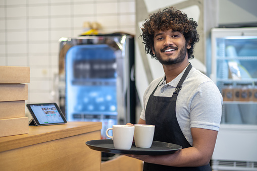 Work, cafe. Young indian man in apron standing with tray of cups smiling looking at camera in cafe