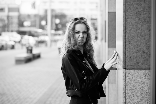 fun sad serious blond woman in leather coat stand on street grimacing looking at camera, monochrome