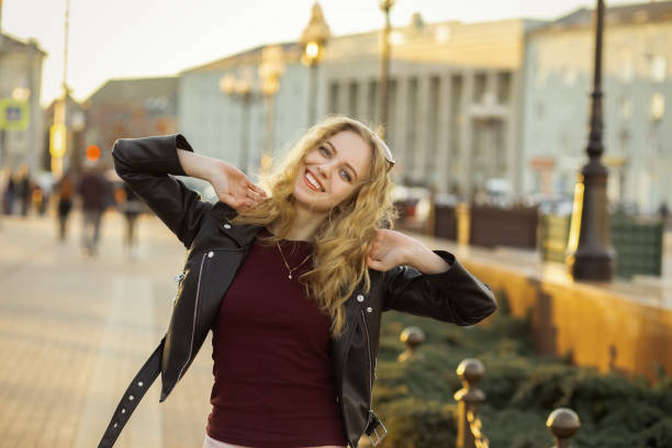 happy blond girl in leather coat looking at camera on city street smiling under sun rays toned image