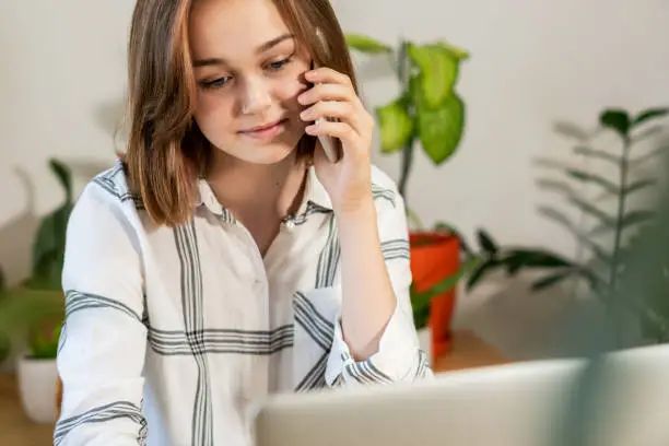 Photo of Young woman working on laptop at home. Happy pretty manager talking on mobile phone with client at home office.