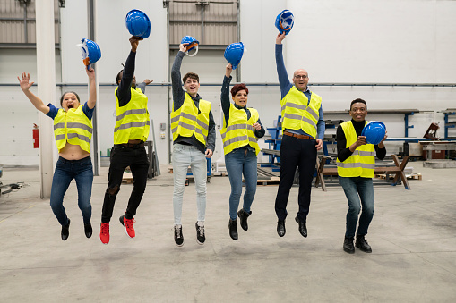 Group of multiracial multigenerational workers taking off helmet inside warehouse and jumping happily, -factory concept-