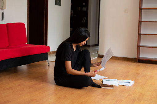 Latin woman of average age of 25 years dressed informally is on the wooden floor of the living room of her house studying with her notes made on sheets of paper