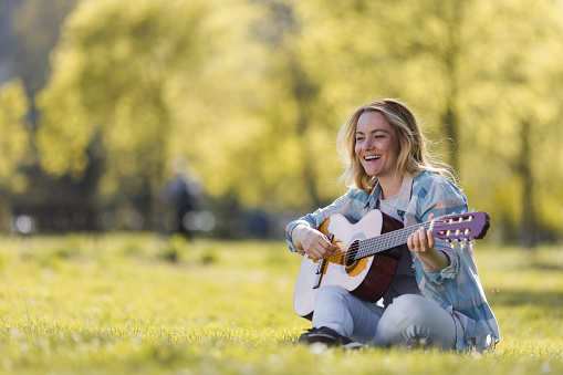 Happy woman sitting on the meadow and playing guitar during spring day