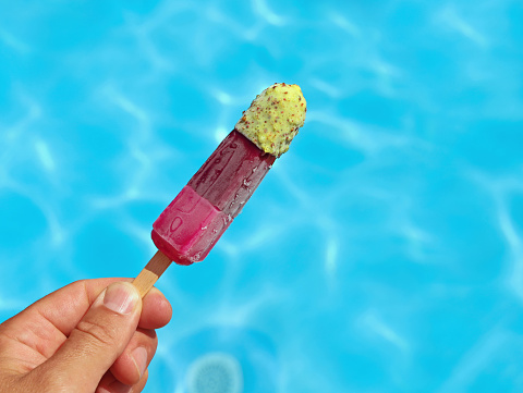 Hand holding fruity colorful popsicles or water ice in front of a blue pool water surface, help against summer heat concept image.