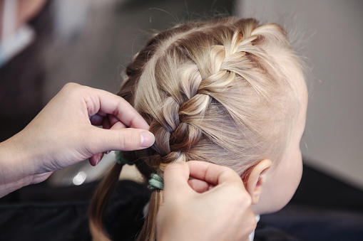 Barber woman braids pigtails for cute little blond girl child, makes fashionable pretty hairstyle in barbershop. Hairdresser makes hairdo for young baby in barber shop. Concept hairstyle and beauty