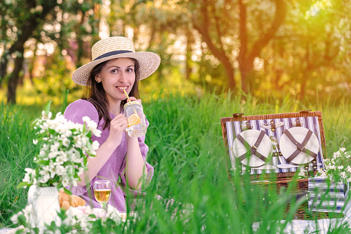 A pretty young woman in a straw hat drinks lemonade at a picnic during sunset