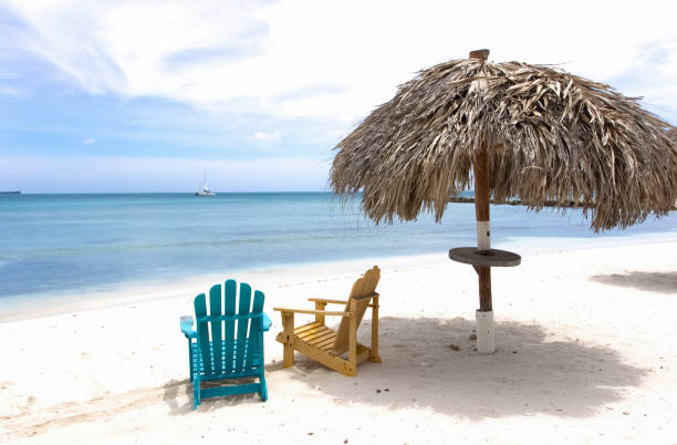 Two wooden beach chairs under thatched roof, Aruba Two wooden beach chairs under thatched roof, Aruba thatched roof hut straw grass hut stock pictures, royalty-free photos & images