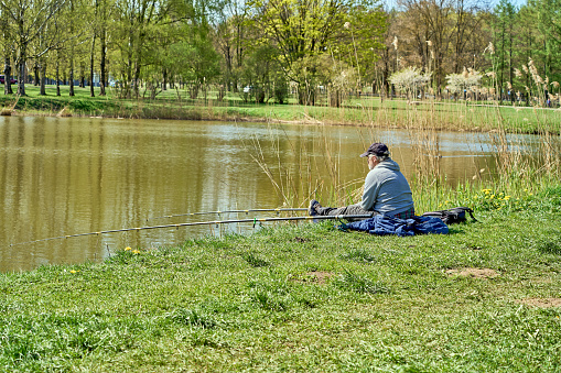 Riga, Latvia - May 8, 2022: A fisherman sits with a fishing rod on the shore of a lake in a public park. Green grass around. View from back.