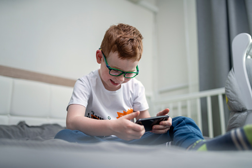 Cute joyful red-haired boy playing games on the smartphone in bedroom