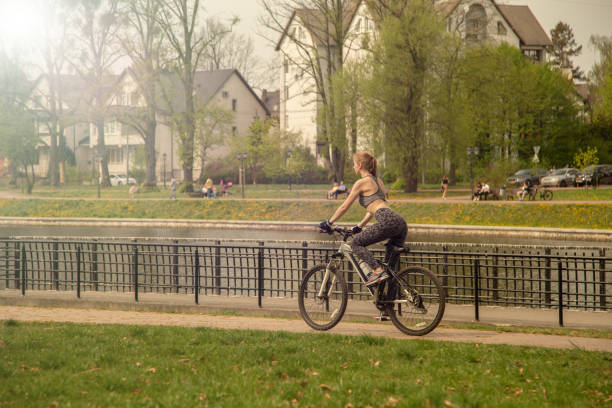 sports slim young blond woman move on bike in park in sun rays, rear side view