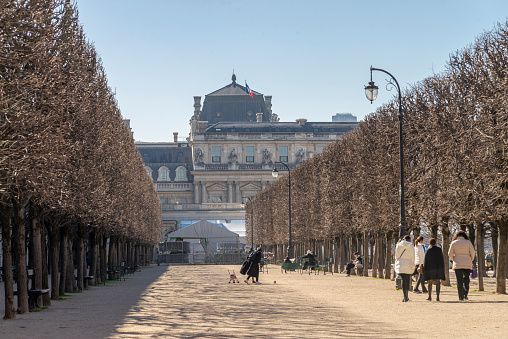 People walking on a gravel footpath near lined trees during a winter day in the Palais Royal Gardens at Paris city, France. The Palais Royal (Royale Palace) at the background.