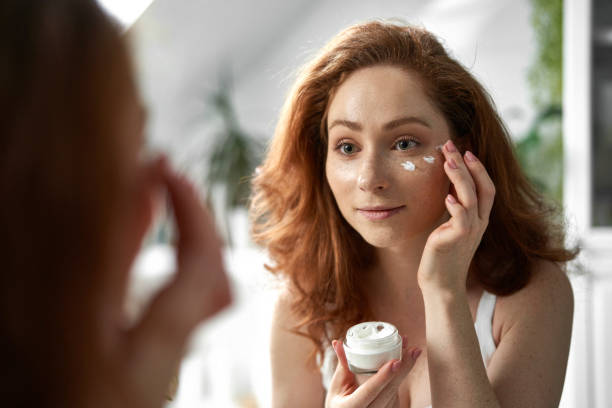 Young caucasian redhead woman smiling and applying face cream stock photo