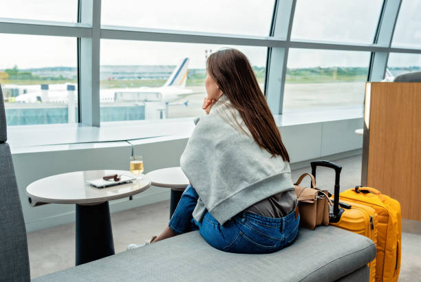 Young woman with yellow suitcase sitting in airport business lounge waiting for plane departure enjoying glass of white vine looking at window at plane. stock photo