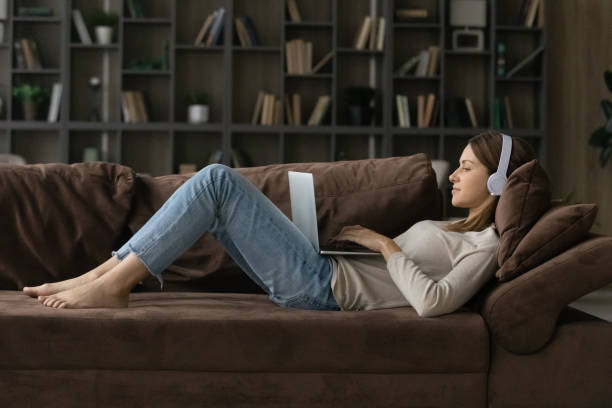 mujer acostada en un cómodo sofá usa auriculares usando computadora portátil - voip telecommunications equipment gratis freedom fotografías e imágenes de stock