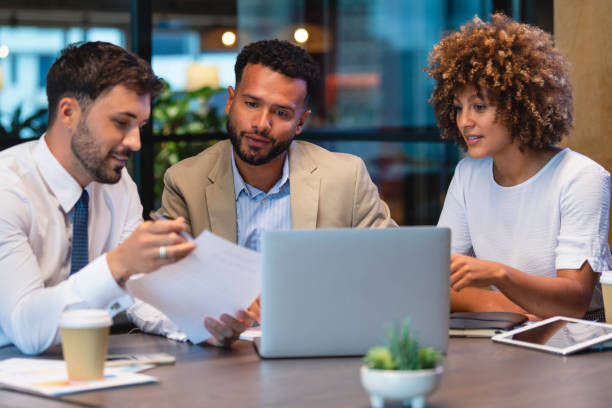 Three business people meeting and looking at a laptop and a document. Three business people meeting and looking at a laptop and a document. There is paperwork and other technology on the table, formal business wear. Homebuying stock pictures, royalty-free photos & images