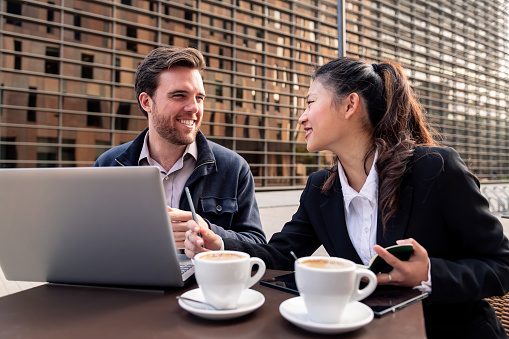 asian woman and caucasian man working with a laptop computer on the terrace of a coffee shop in the financial district, concept of entrepreneurship and business
