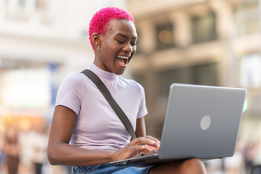 Stylish young afro woman laughing while using a laptop on the street
