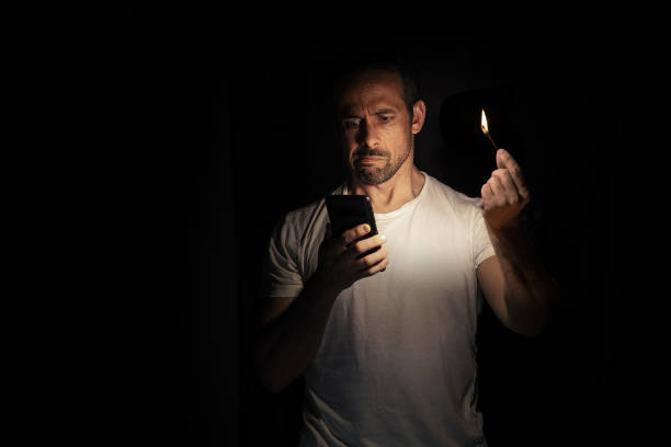 Man looking at his cell phone with a match in the dark. Blackout concept. Selective focus. Man looking at his cell phone with a match in the dark. Blackout concept. Selective focus. blackout stock pictures, royalty-free photos & images