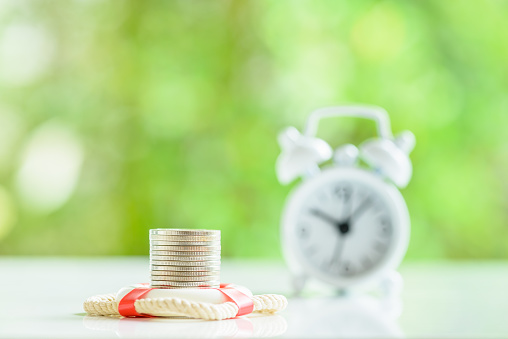 Long-term money saving and wealth protection over time, financial concept : Coins in a red lifebuoy, a white alarm clock on a table. The image depicting securing value of money by applying an insurance, insured assets for safe and secured future.