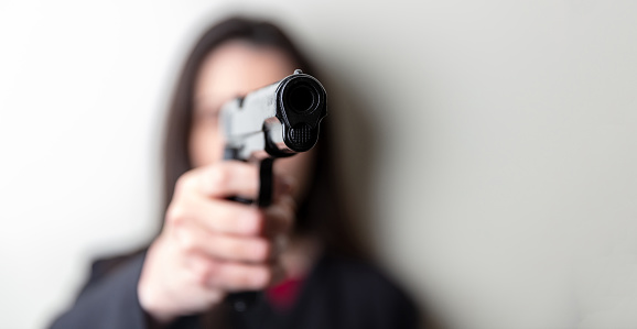 Woman holding a gun in hand pointing at camera. Female secret police agent, killer or spy dark long hair, copy space. Selective focus