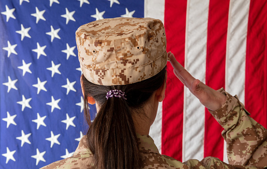 Female US Army Soldier standing in front of an American flag and saluting. Woman in military uniform rear view