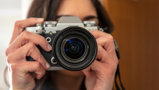 Young woman holding photo camera is shooting your picture. Girl photographer focus at you, close up view