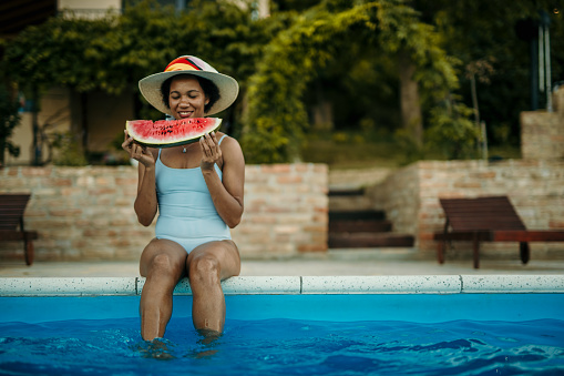 Loving and cheerful young adult, African American, woman, sitting by the pool with her feet in the water, enjoying watermelon. She's wearing a cute blue stripped swimming suit with a summer ha on her head. She's radiating enjoyment and happiness