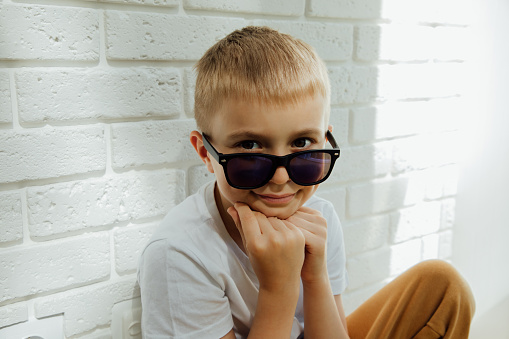 A boy in sunglasses sits leaning against a brick wall and smiles. Portrait of a boy 6-7 years old