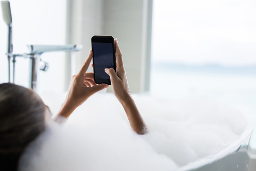 Close up of unrecognizable relaxed woman text messaging on smart phone in her bubble bath in a bathroom.