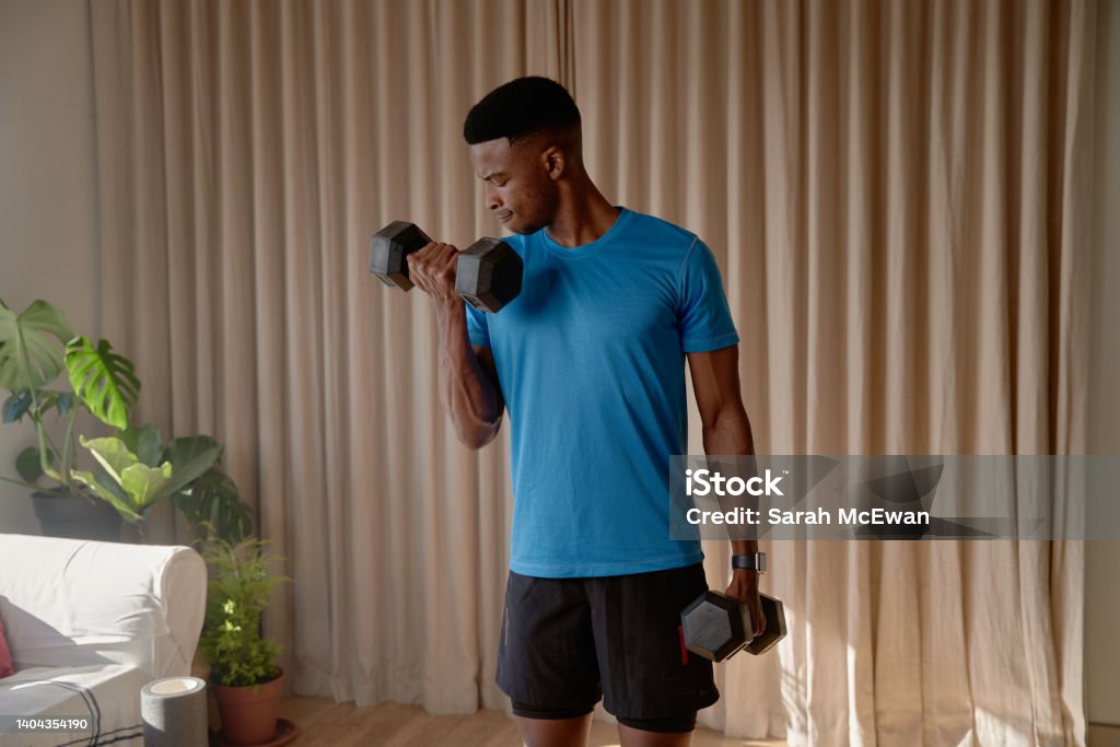 Young African American black male working out in living room at home doing a bicep curl with heavy dumbbell weights. Keeping fit and healthy with strength training Young African American black male working out in living room at home doing a bicep curl with heavy dumbbell weights. Keeping fit and healthy with strength training. High quality photo Bicep Stock Photo