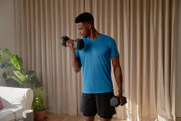 Photo of Young African American black male working out in living room at home doing a bicep curl with heavy dumbbell weights. Keeping fit and healthy with strength training