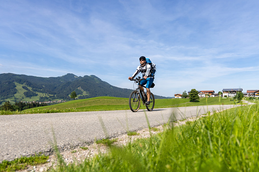 A cyclist with a backpack on a mountain bike is riding through beautiful nature in the southern part of Bavaria, Germany. Sunny weather, green gras and sunshine with a mountain in the background.