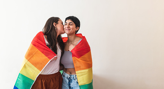 Authentic shot of happy lesbian couple wrapped in rainbow flag and giving a kiss, copy space on the right - lesbian couple at home enjoying life together