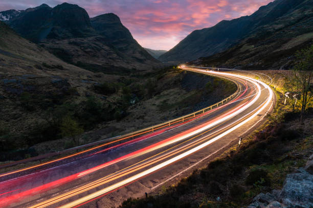 senderos ligeros para automóviles en una carretera sinuosa a través de las tierras altas cerca de glencoe en escocia al atardecer - curve driving winding road landscape fotografías e imágenes de stock