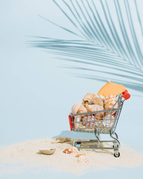 istock Shopping cart filled with seashells and parasol placed on sand on bright blue background decorate with palm leaf shadow. Minimal summer concept. Creative tropical vacation layout. 1404367348