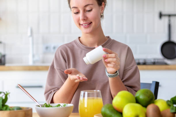 vitamine, integratori alimentari, concetto di stile di vita sano. giovane donna che fa colazione e prende la vitamina seduta a casa in cucina, sorridente amichevole - vitamin pill nutritional supplement pill healthcare and medicine foto e immagini stock