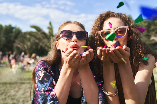 Two young caucasian women blowing confetti towards camera on music festival