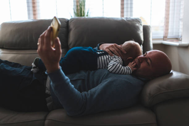 Man lying on the sofa with his son and using smartphone stock photo