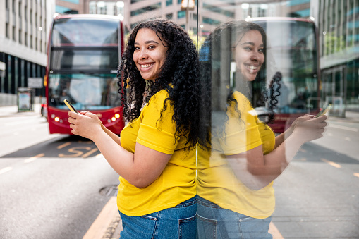 Happy woman with curly hair and mixed race look using her smartphone, smiling at the camera and waiting at bus stop in the city - Lifestyle and transport in Birmingham